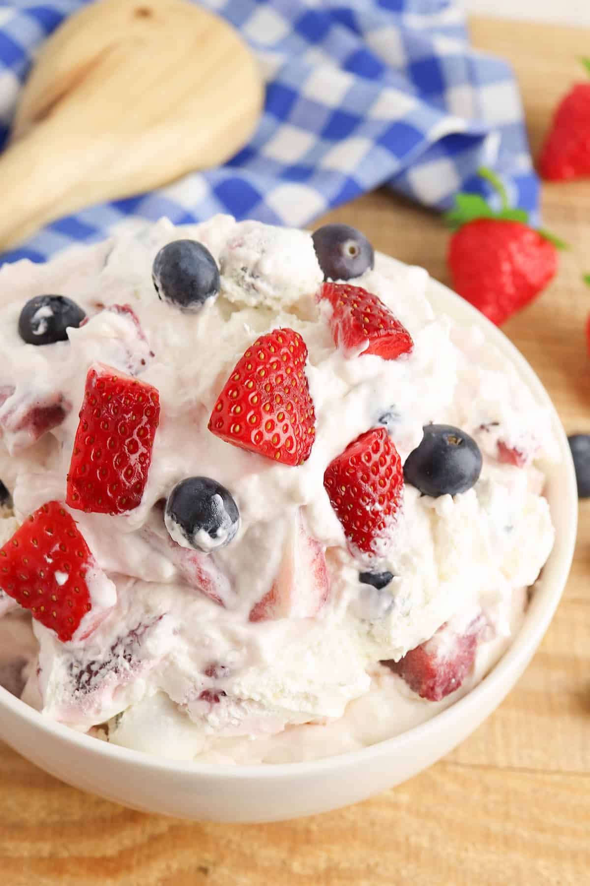 A bowl of creamy Berry Cheesecake Salad, topped with sliced strawberries and whole blueberries, sits on a wooden surface with a blue checkered cloth and whole strawberries in the background.