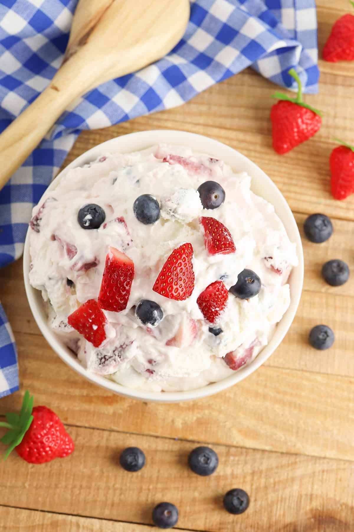 A bowl of Berry Cheesecake Salad, with whipped cream mixed with strawberries and blueberries, sits on a wooden table, surrounded by fresh berries and a blue checkered cloth.