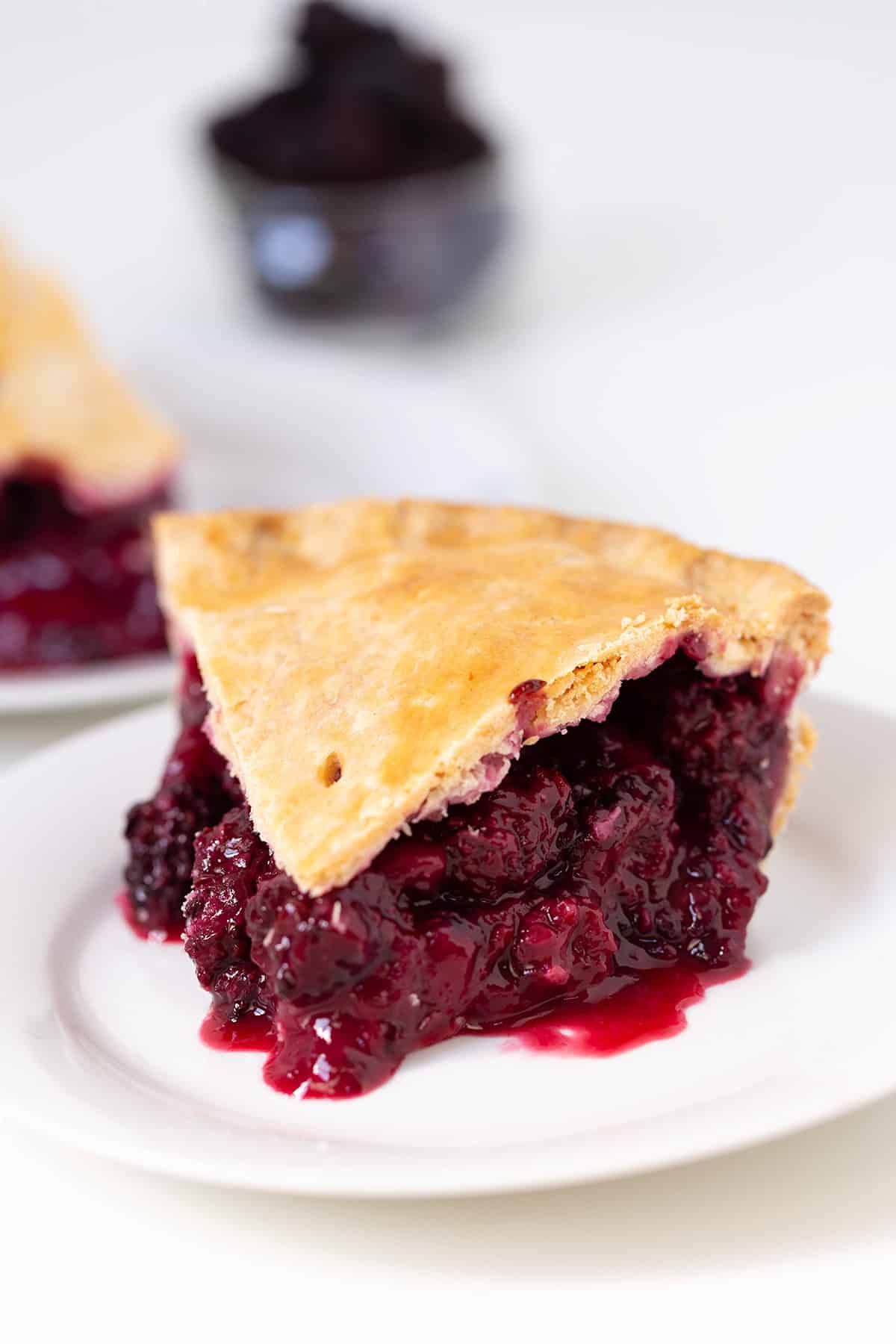 A slice of blackberry pie with a golden crust sits on a white plate, its rich berry filling and sauce dripping out, capturing the essence of homemade blackberry pie perfection.