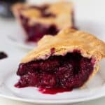A close-up of a slice of blackberry pie with a golden crust and thick, juicy berry filling on a white plate.