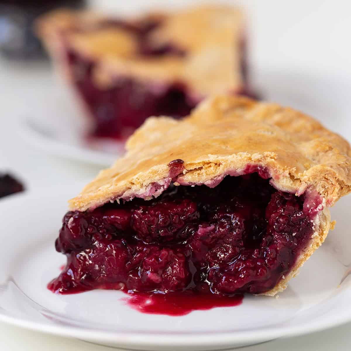 A close-up of a slice of blackberry pie with a golden crust and thick, juicy berry filling on a white plate.
