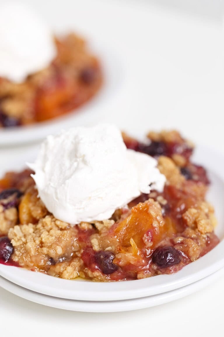 A serving of Blueberry Peach Crisp topped with a scoop of vanilla ice cream on a white plate.