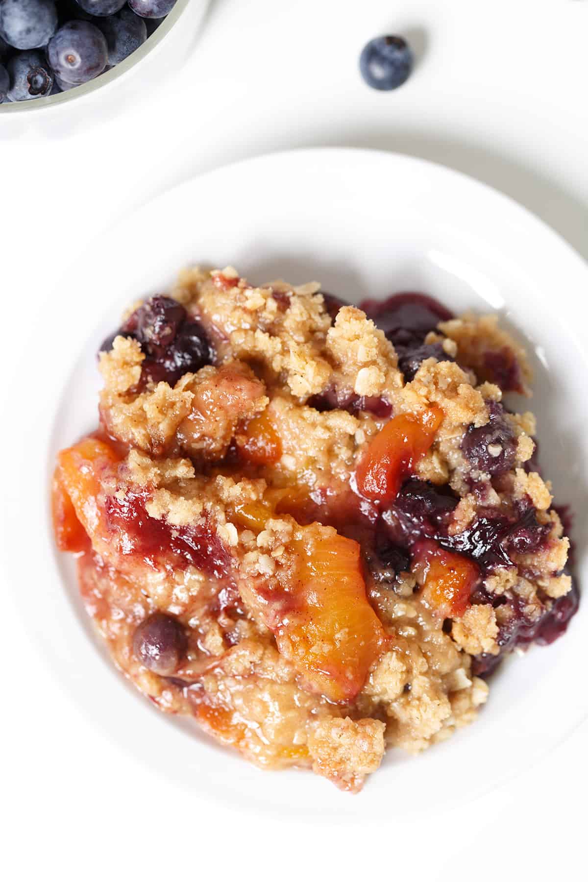 A close-up of a blueberry peach dessert featuring a serving of fruit crumble with peaches and blueberries on a white plate, with a bowl of blueberries and a few loose berries in the background.