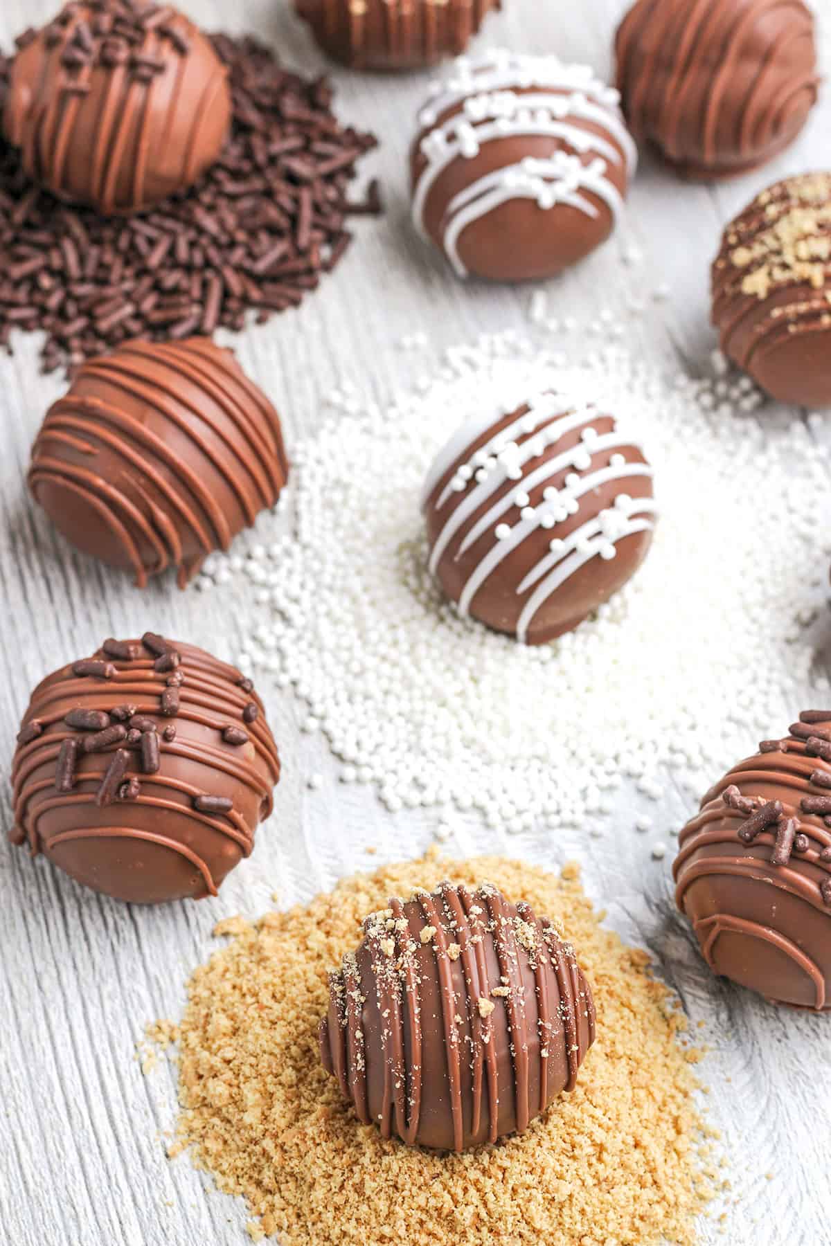 Assorted cheesecake truffles with decorative drizzles and toppings are arranged on small piles of sprinkles and crumbs on a light wooden surface.