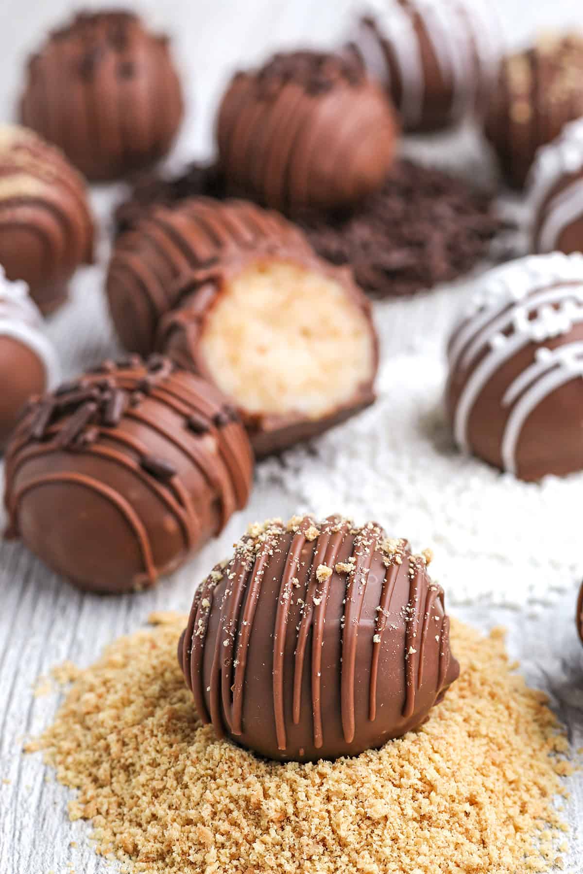 Assorted cheesecake truffles, some drizzled with chocolate or white icing, arranged on a white wooden surface with one truffle cut open and crumbs scattered around.