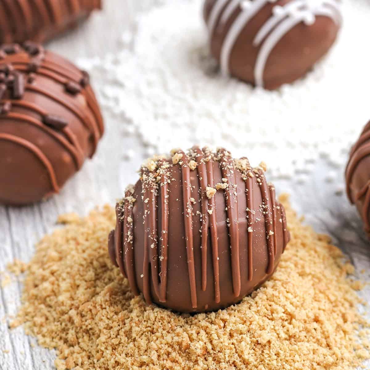 A close-up of cheesecake truffles, one resting on a bed of crushed crumbs, topped with drizzled chocolate and sprinkled crumbs; others are in the background.