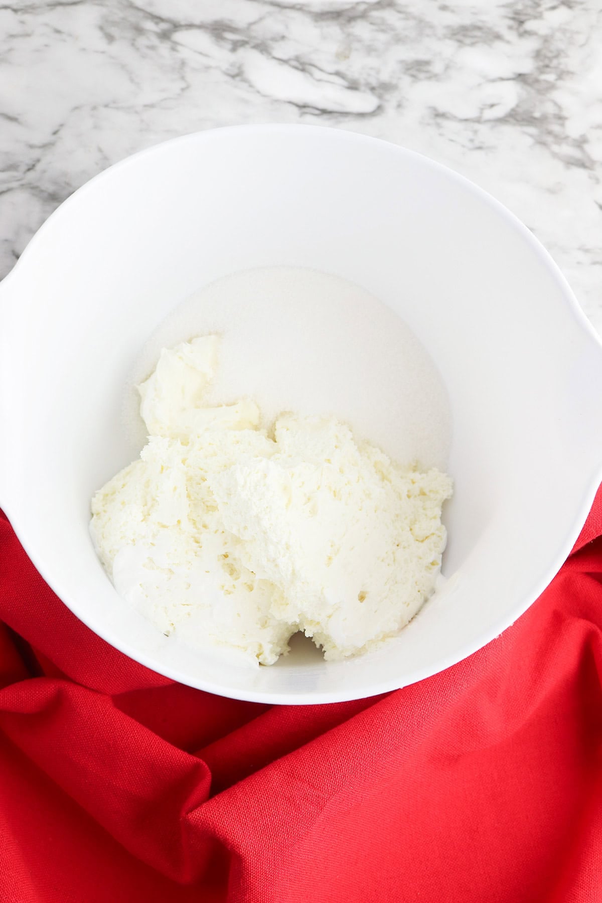 A white mixing bowl containing cream cheese and granulated sugar sits on a marble countertop with a red cloth underneath, ready to be transformed into a delicious Berry Cheesecake Salad.