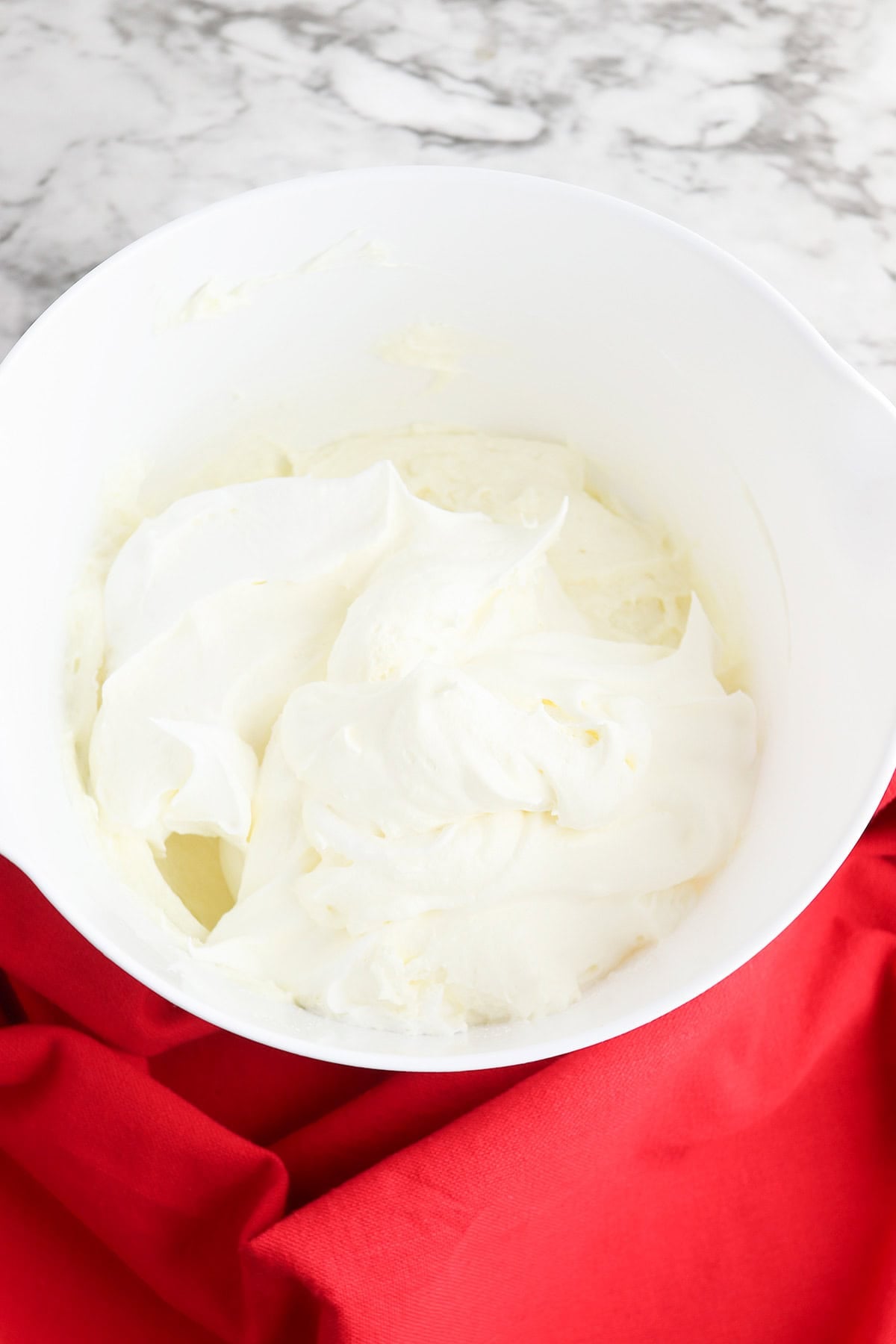 A white mixing bowl filled with whipped cream for Berry Cheesecake Salad sits on a red cloth atop a marble surface.