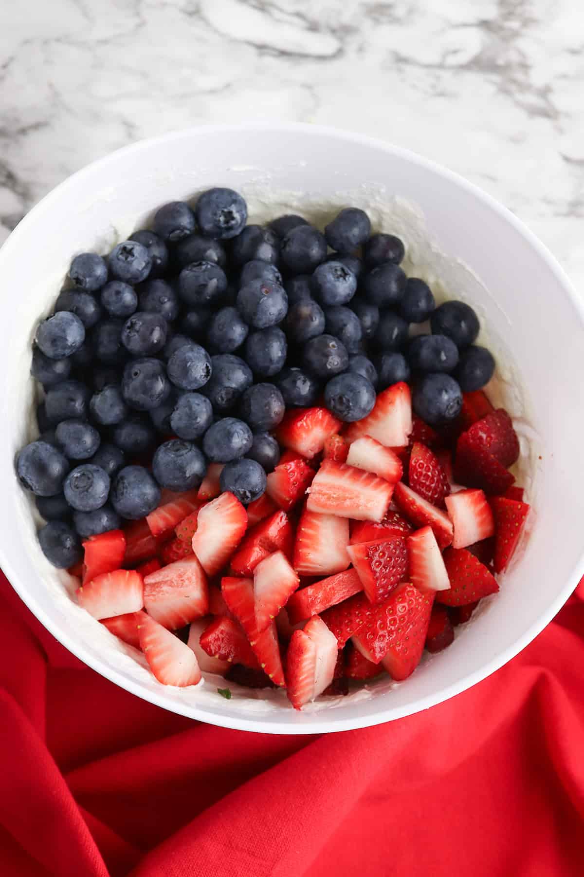 A white bowl filled with fresh berry salad—blueberries and chopped strawberries—sits on a red cloth with a marble countertop in the background.
