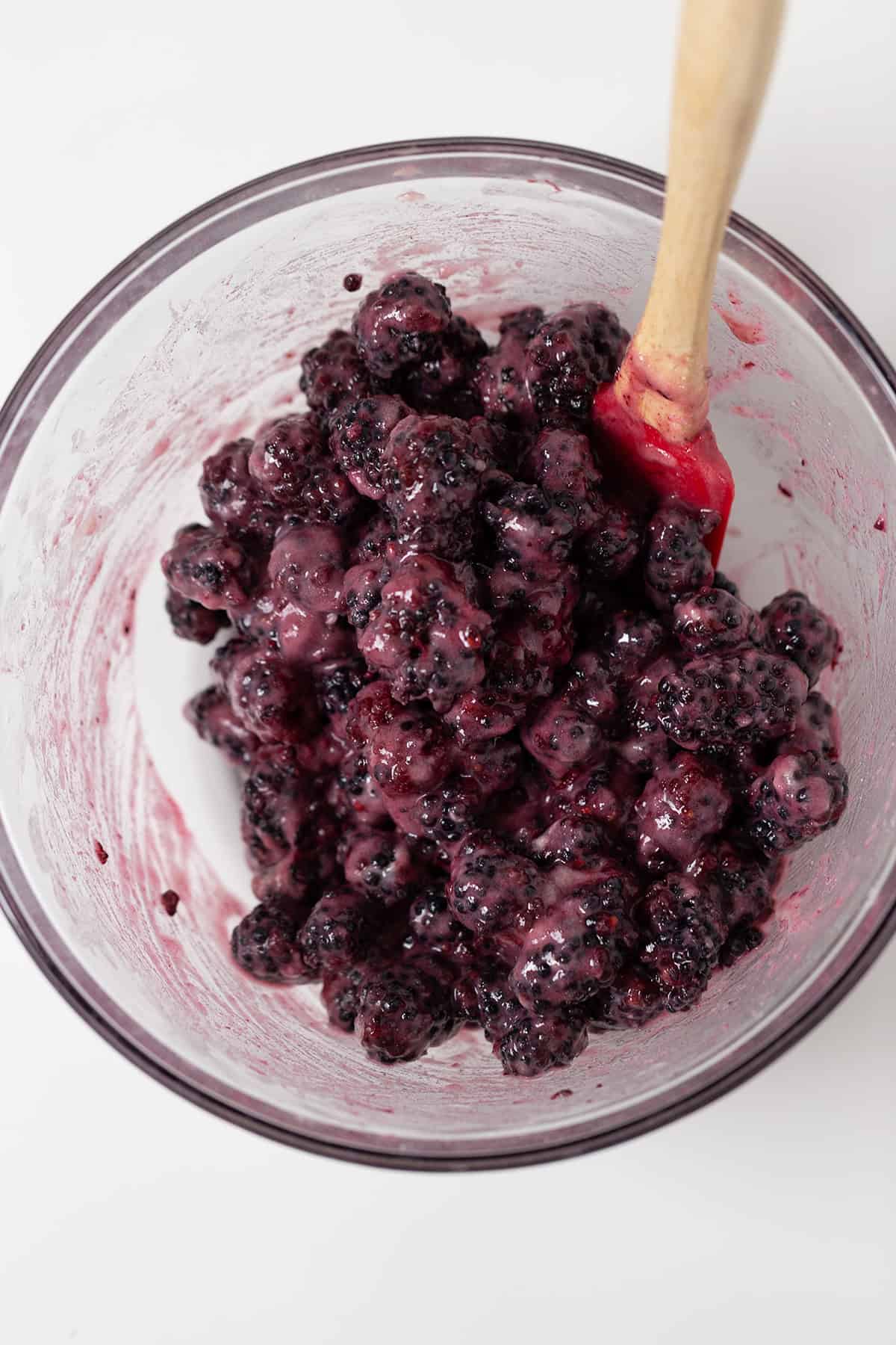 A glass bowl filled with blackberries coated in a creamy mixture, this tempting blackberry dessert is finished off with a red spatula resting inside.