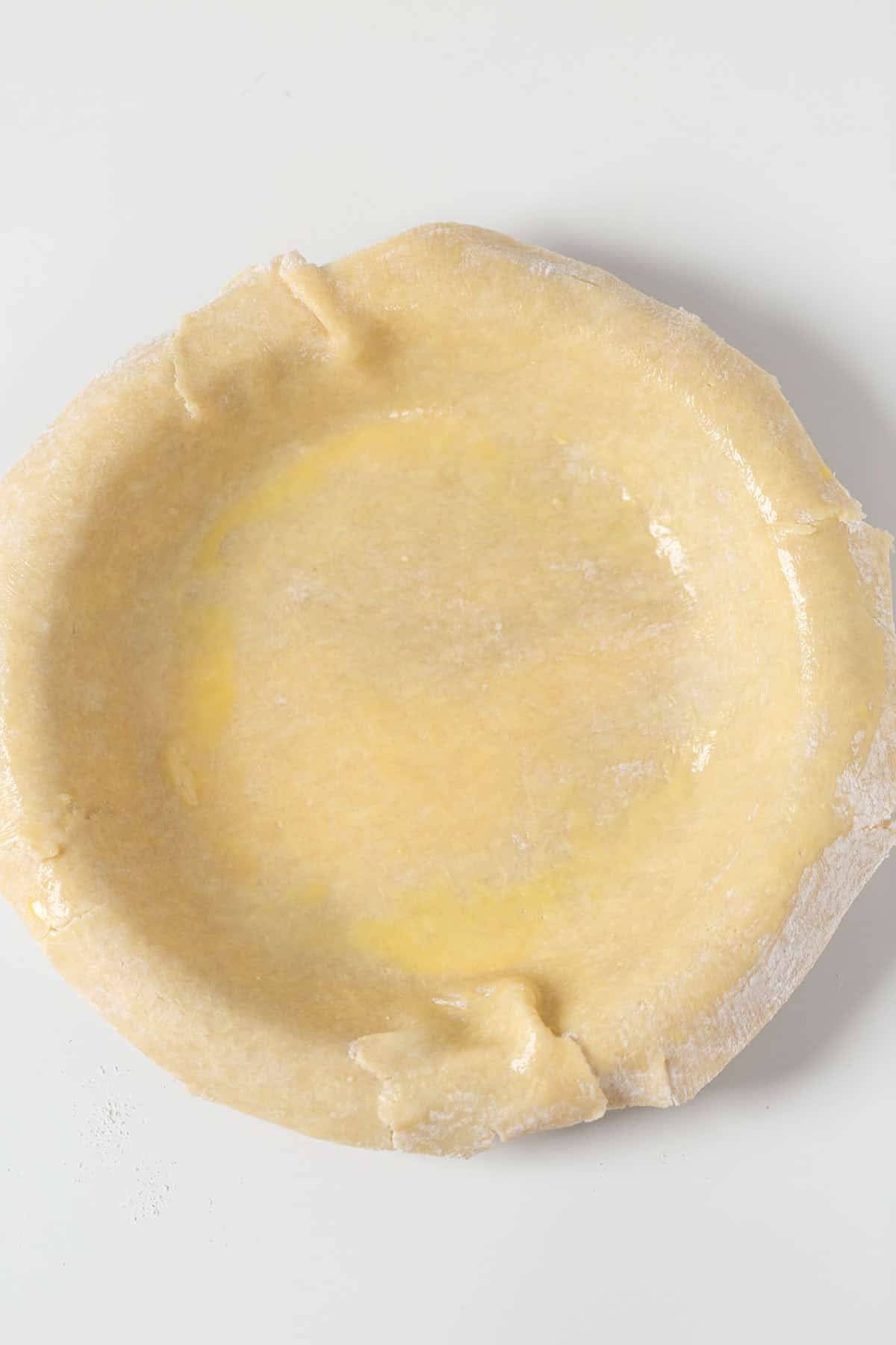 Unbaked pie crust in a pie dish on a white surface, edges slightly uneven and dusted with flour&mdash;perfect for starting your favorite homemade Blackberry Pie recipe.