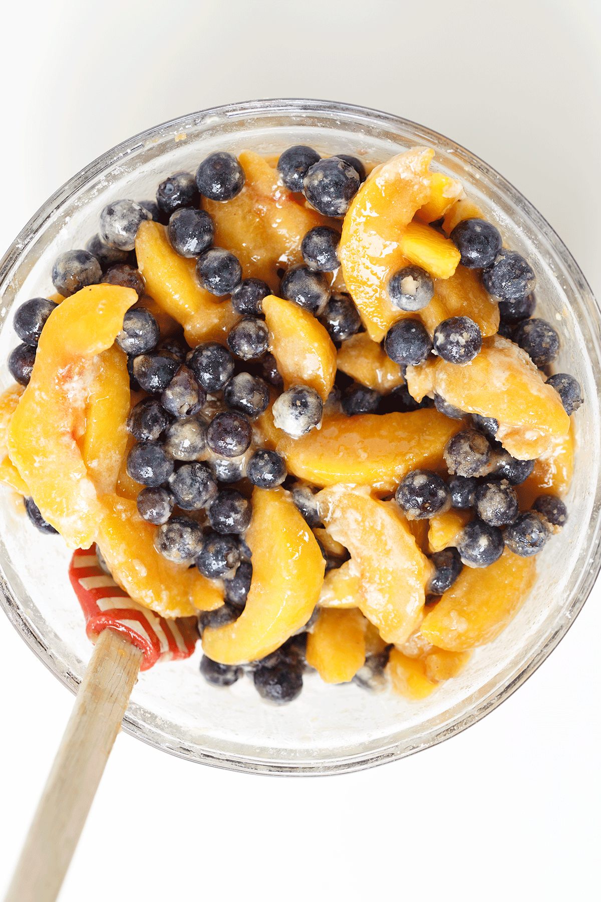 A glass bowl filled with sliced peaches and blueberries, mixed with sugar for a fresh blueberry peach dessert, with a wooden spoon resting inside.