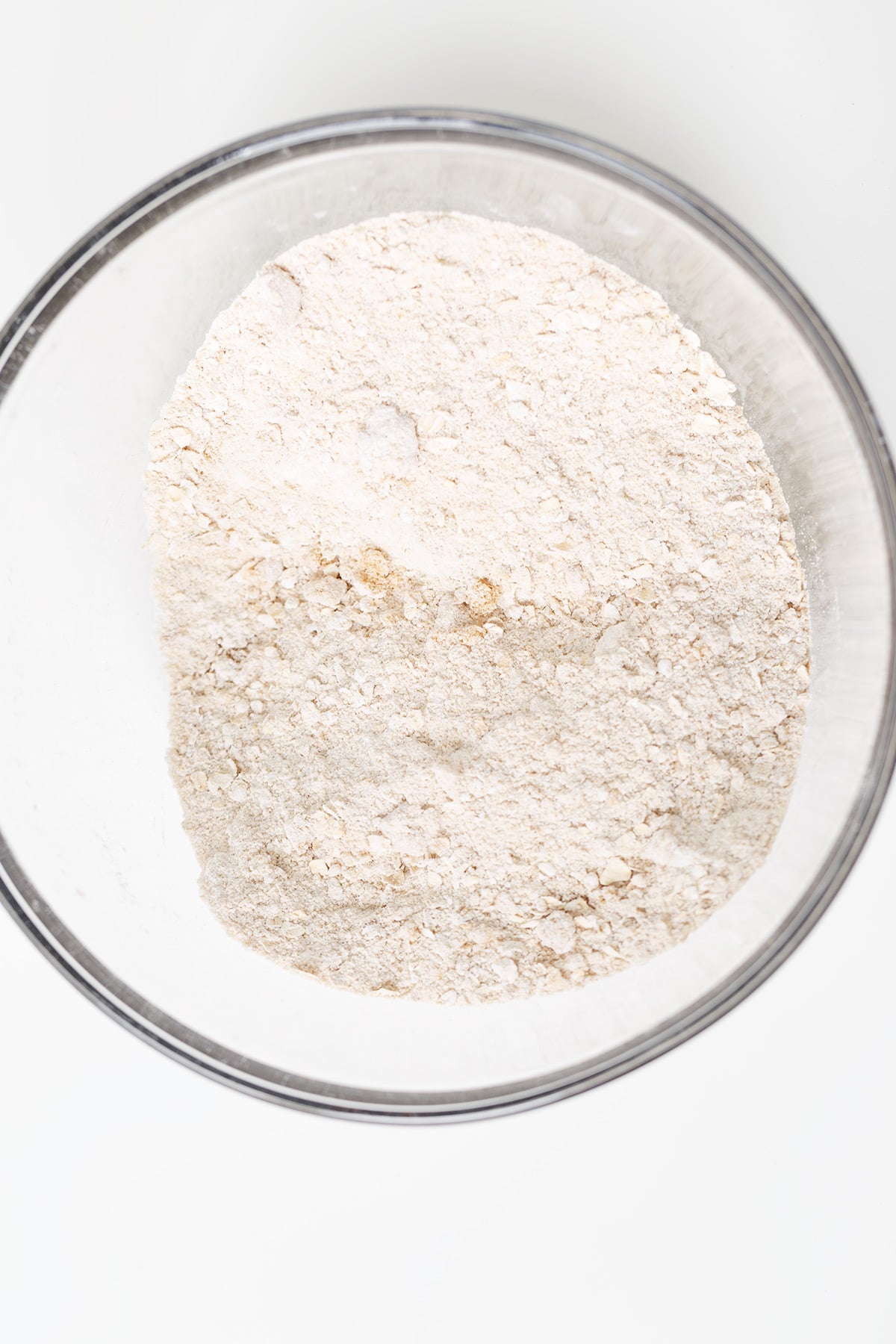 A glass bowl filled with a mixture of dry ingredients, likely flour and oats, perfect for making a blueberry or peach crisp, sits on a white surface.