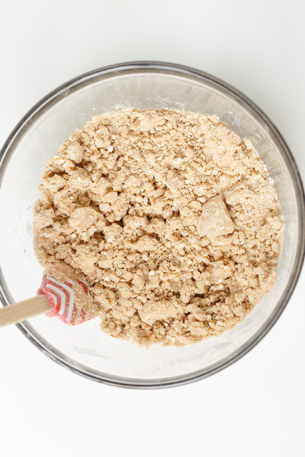 A glass bowl filled with crumbly dough mixture for Peach Crisp and a red and white spatula, viewed from above on a white surface.