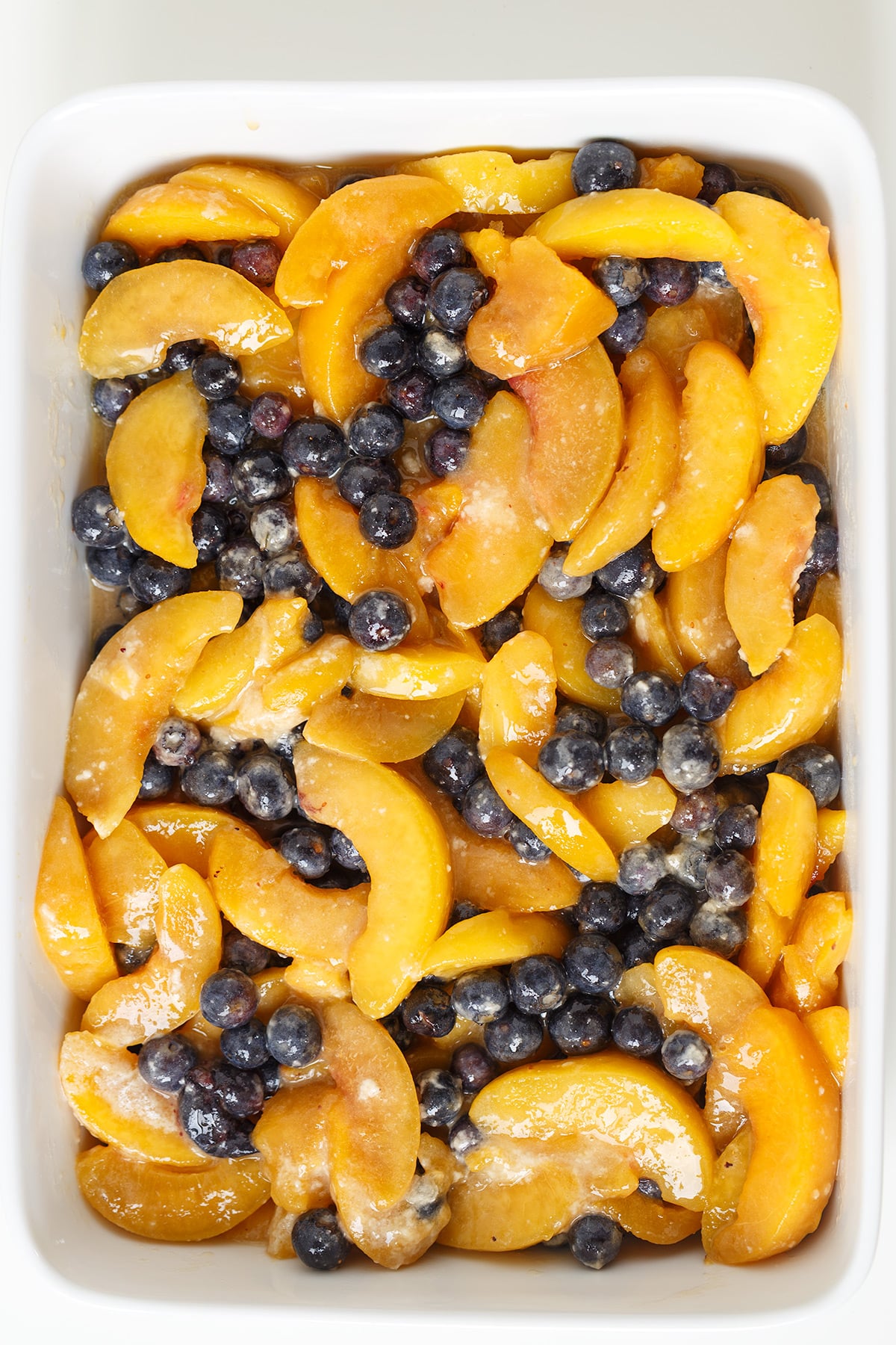 Sliced peaches and blueberries mixed with sugar and butter in a white baking dish, ready for baking into a delicious Blueberry Peach Crisp.