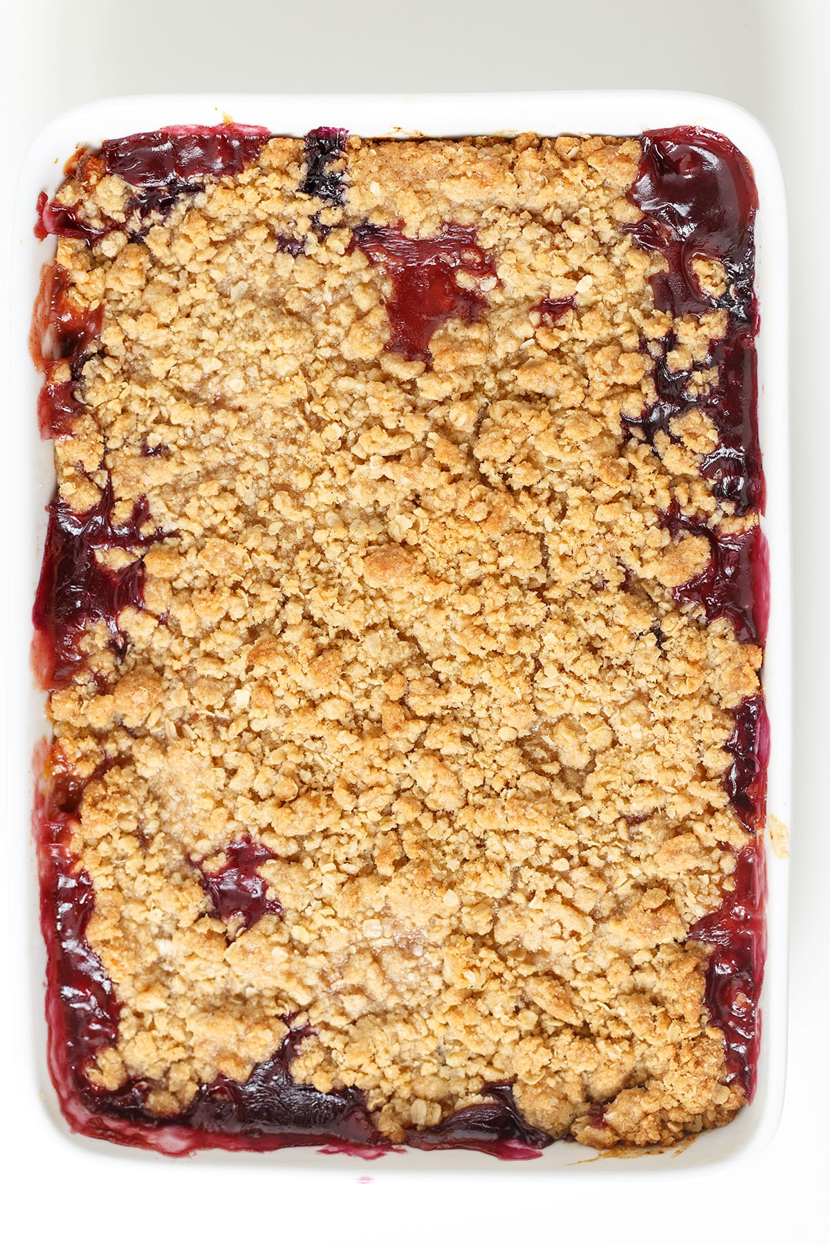 A rectangular dish filled with Blueberry Peach Crisp, featuring a golden brown oat topping and visible dark red fruit filling along the edges.