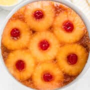 A classic pineapple upside down cake made with cake mix, topped with caramelized pineapple rings and maraschino cherries on a white plate, with extra pineapple slices in a bowl nearby.
