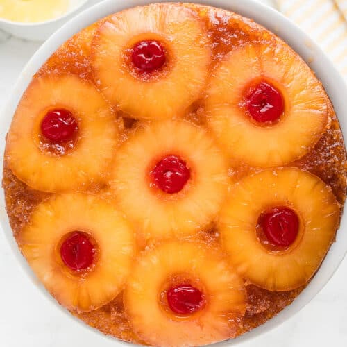 A classic pineapple upside down cake made with cake mix, topped with caramelized pineapple rings and maraschino cherries on a white plate, with extra pineapple slices in a bowl nearby.