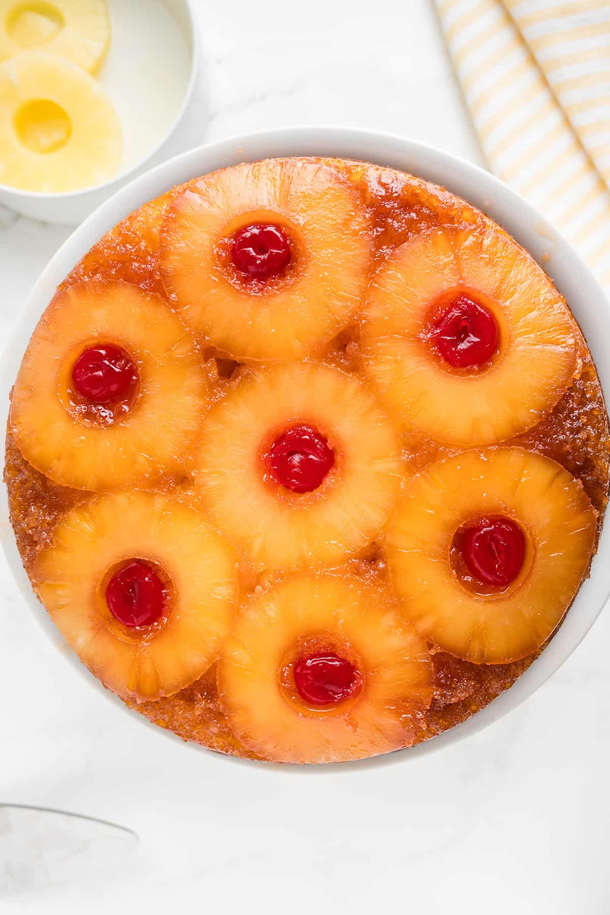 A classic pineapple upside down cake made with cake mix, topped with caramelized pineapple rings and maraschino cherries on a white plate, with extra pineapple slices in a bowl nearby.