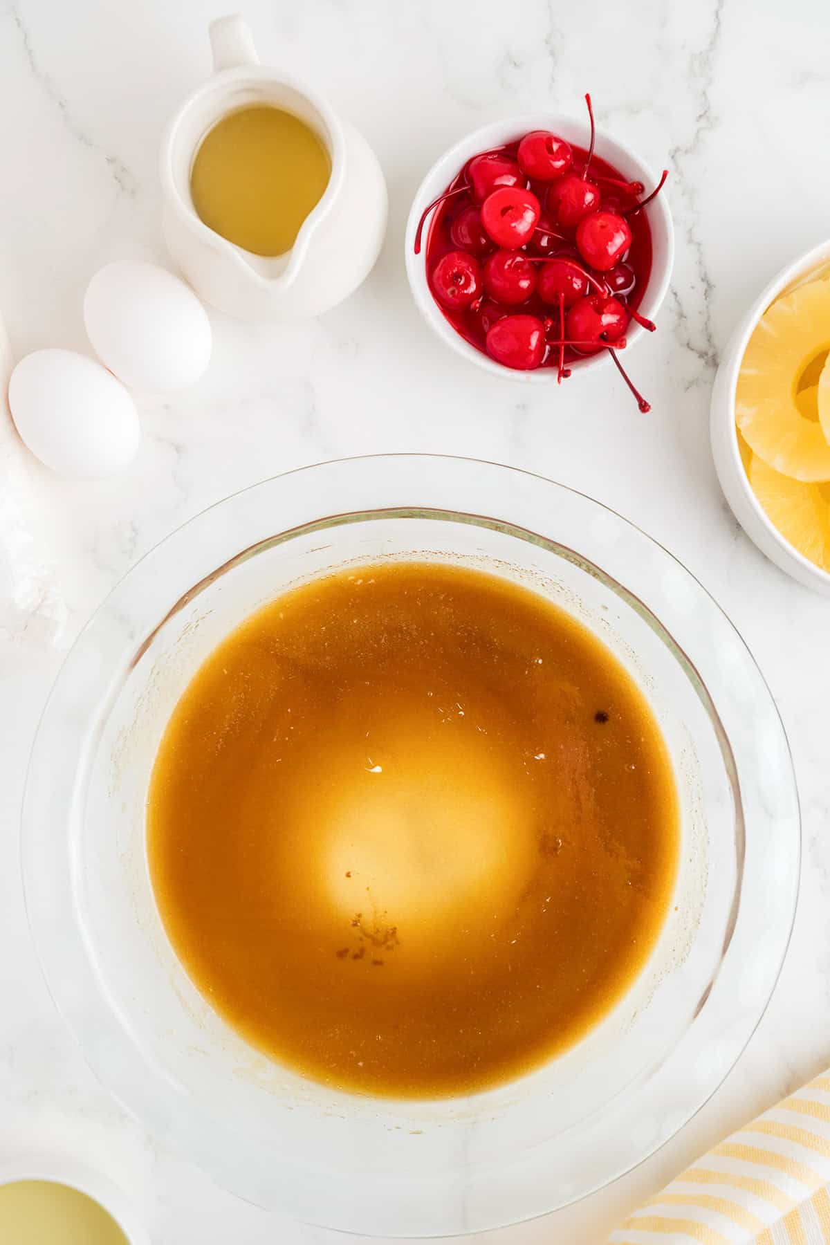 Overhead view of a glass bowl with brown cake batter made from cake mix, surrounded by eggs, a small pitcher of liquid, maraschino cherries, and pineapple rings—perfect for pineapple upside down cake on a marble countertop.