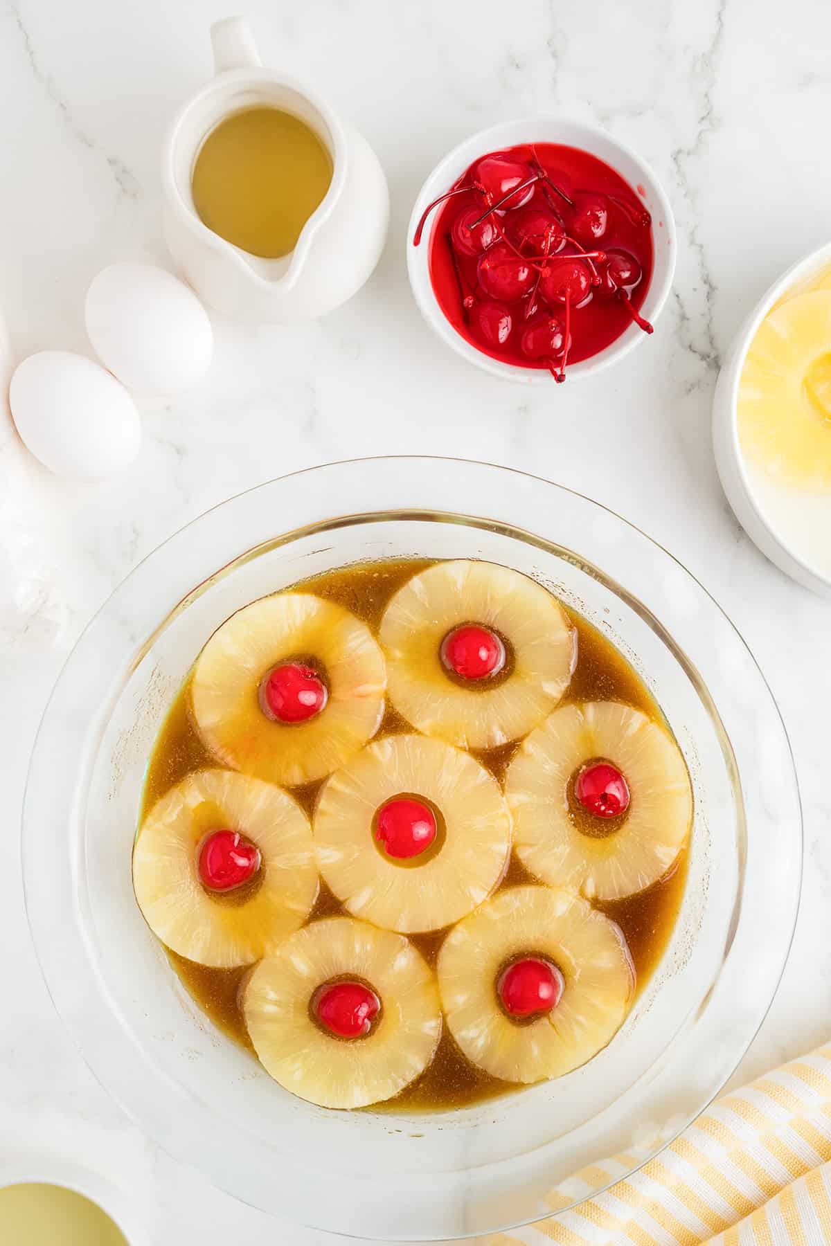 A glass dish with pineapple rings and maraschino cherries in brown sauce, just like a classic pineapple upside down cake, surrounded by eggs, a bowl of cherries, pineapple slices, and a jug of liquid on a white surface.