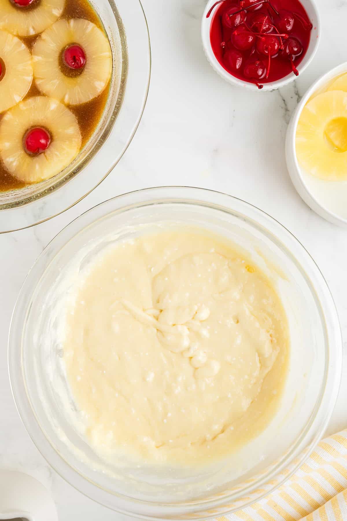 A glass bowl of cake mix sits on a counter beside a pan for pineapple upside down cake, with pineapple rings, cherries, a bowl of maraschino cherries, and a plate with pineapple slices.