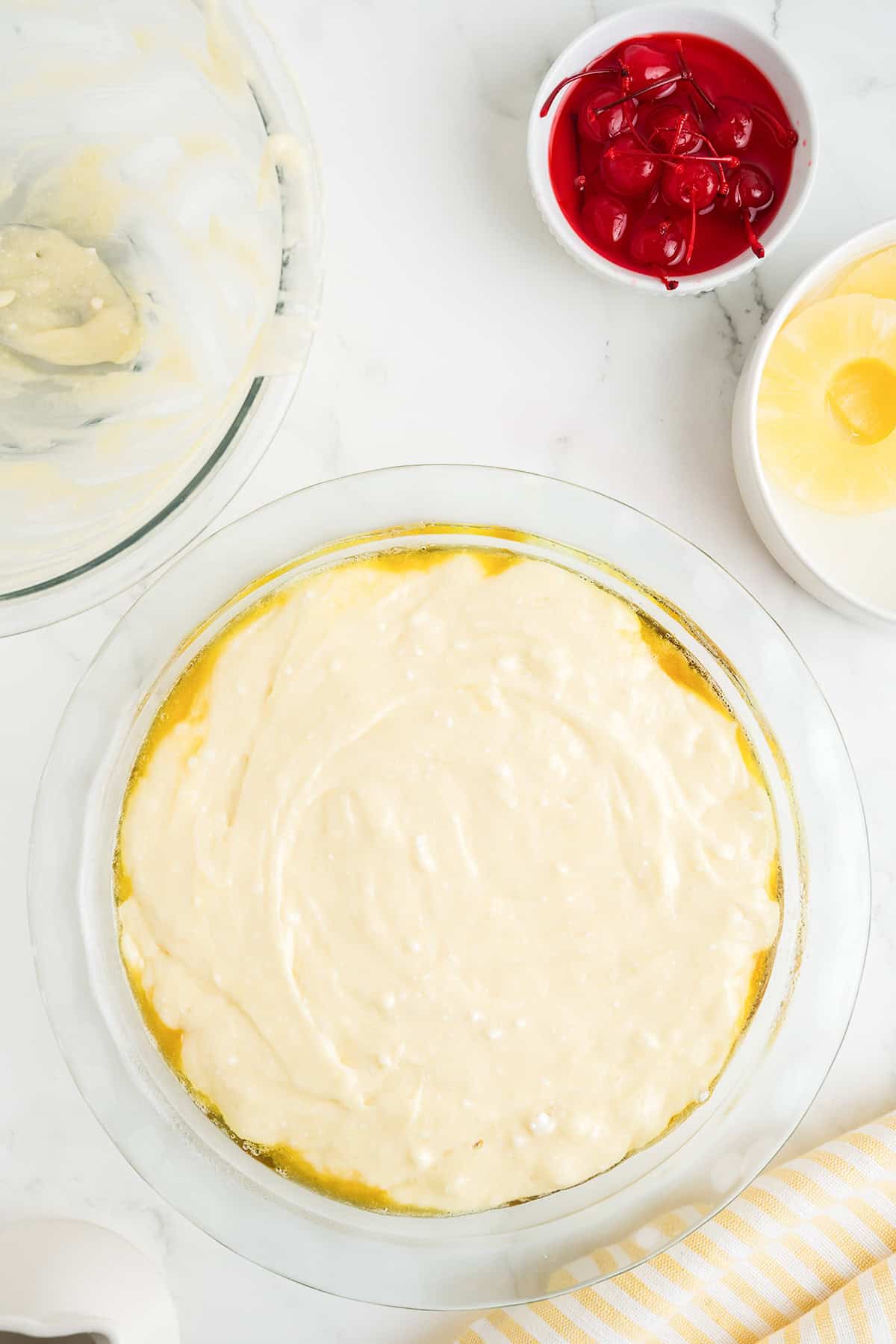 A glass pie dish filled with cake batter sits on a white counter near a bowl of maraschino cherries, pineapple rings, and an empty mixing bowl—everything you need to create a classic pineapple upside down cake using your favorite cake mix.