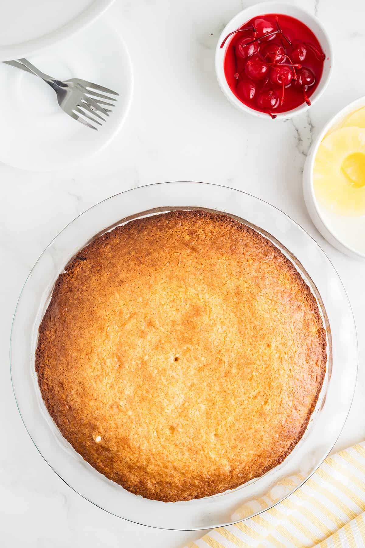 A baked round pineapple upside down cake in a glass pie dish on a white surface, with bowls of maraschino cherries and pineapple slices nearby, plus a plate, fork, and a yellow striped cloth. Perfect for using your favorite cake mix.