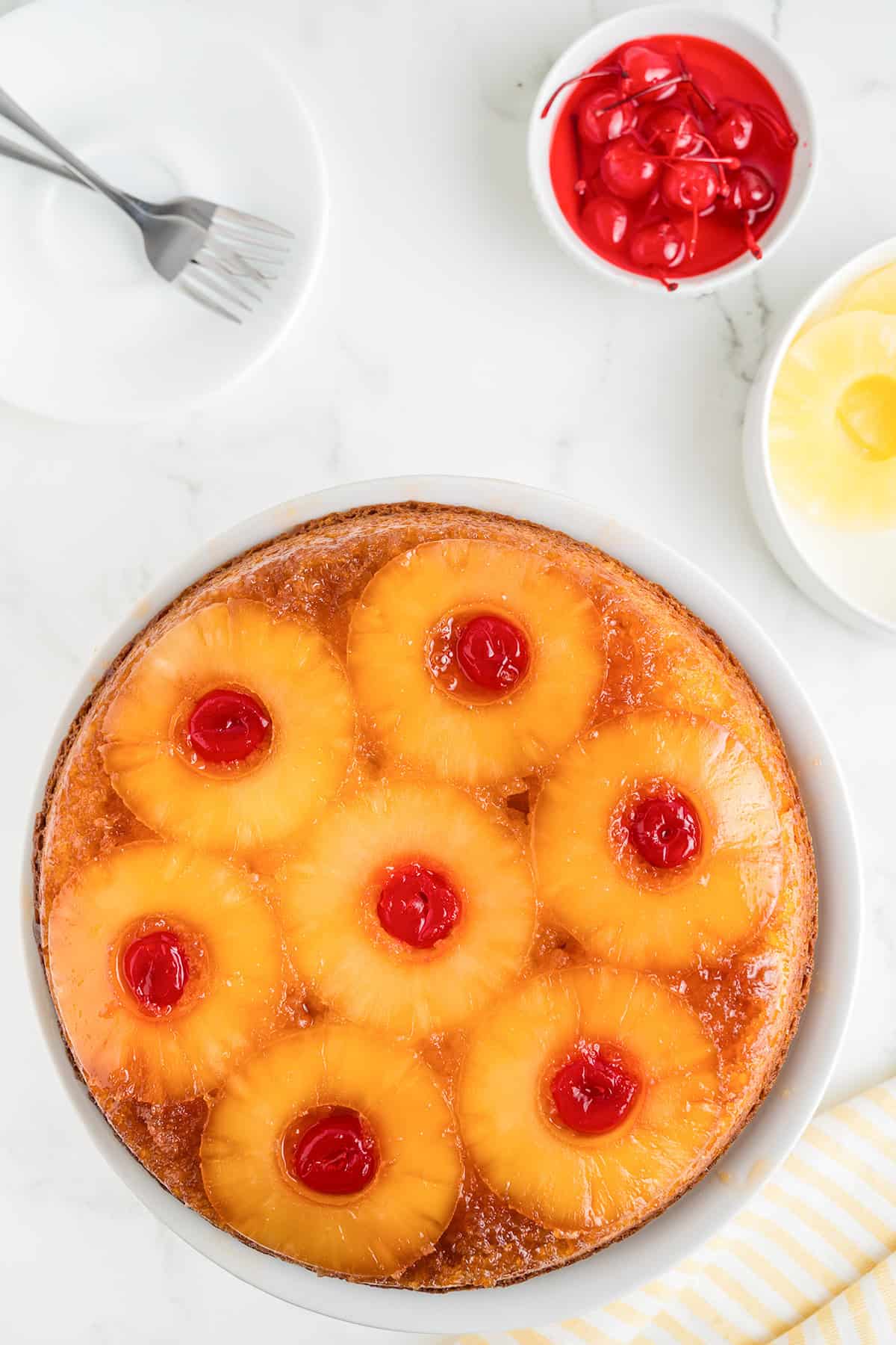 A pineapple upside down cake, made with cake mix and topped with pineapple rings and maraschino cherries, sits on a white plate, surrounded by a bowl of cherries and plates.
