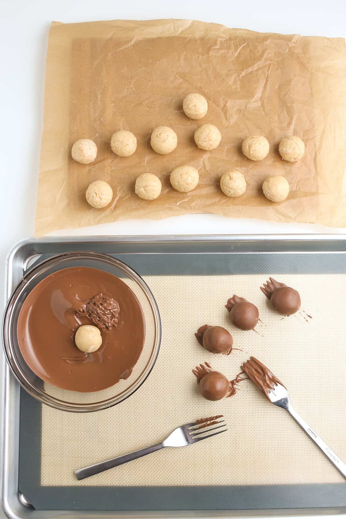 Round peanut butter balls are arranged on parchment paper, with some transformed into decadent truffles by being dipped in melted chocolate and placed on a baking tray lined with a silicone mat.
