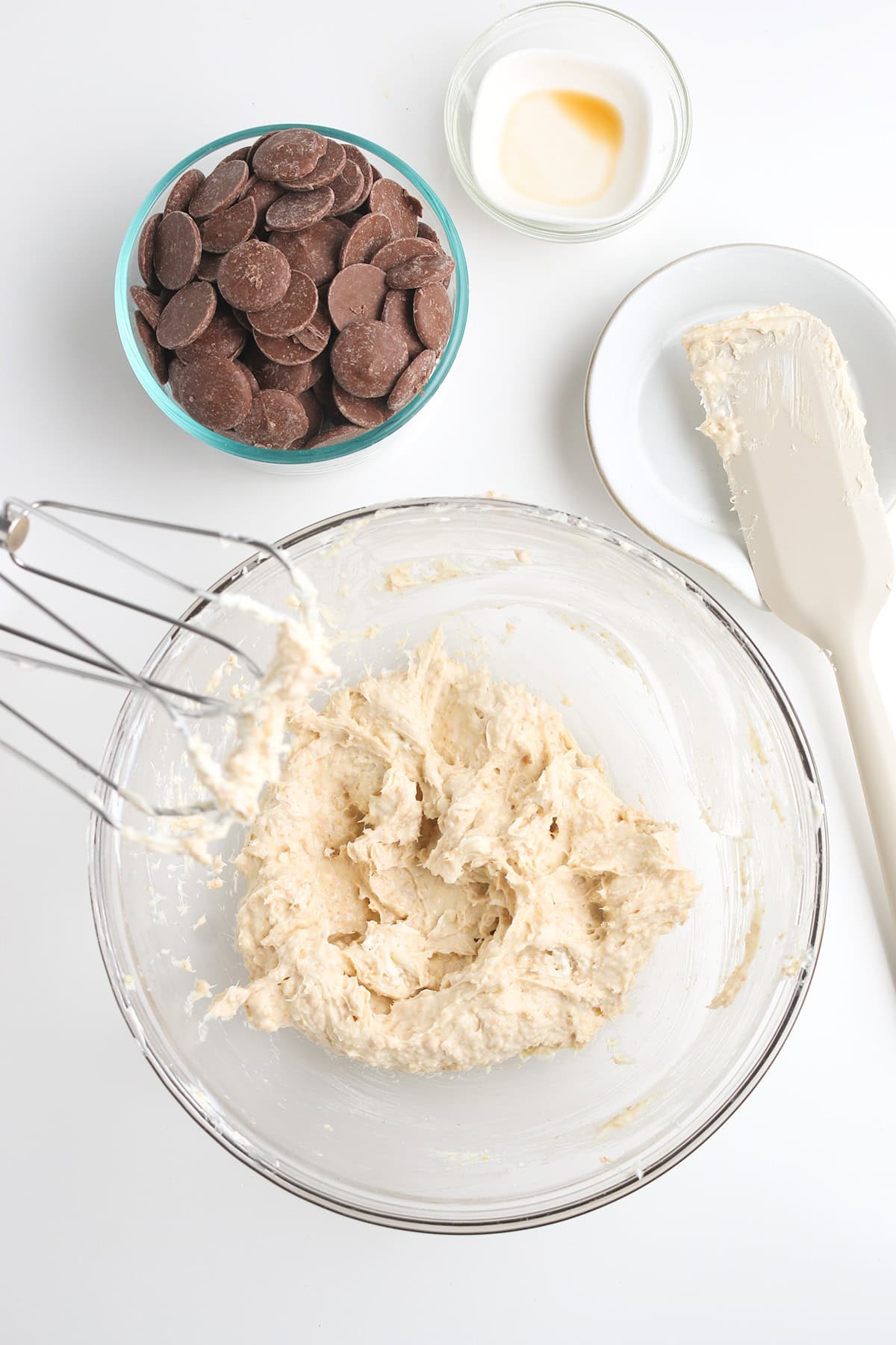 A glass bowl with cookie dough, a metal whisk, a bowl of chocolate wafers, a small dish of vanilla, and a plate with a spatula covered in dough set on a white surface&mdash;perfect for making cheesecake truffles.