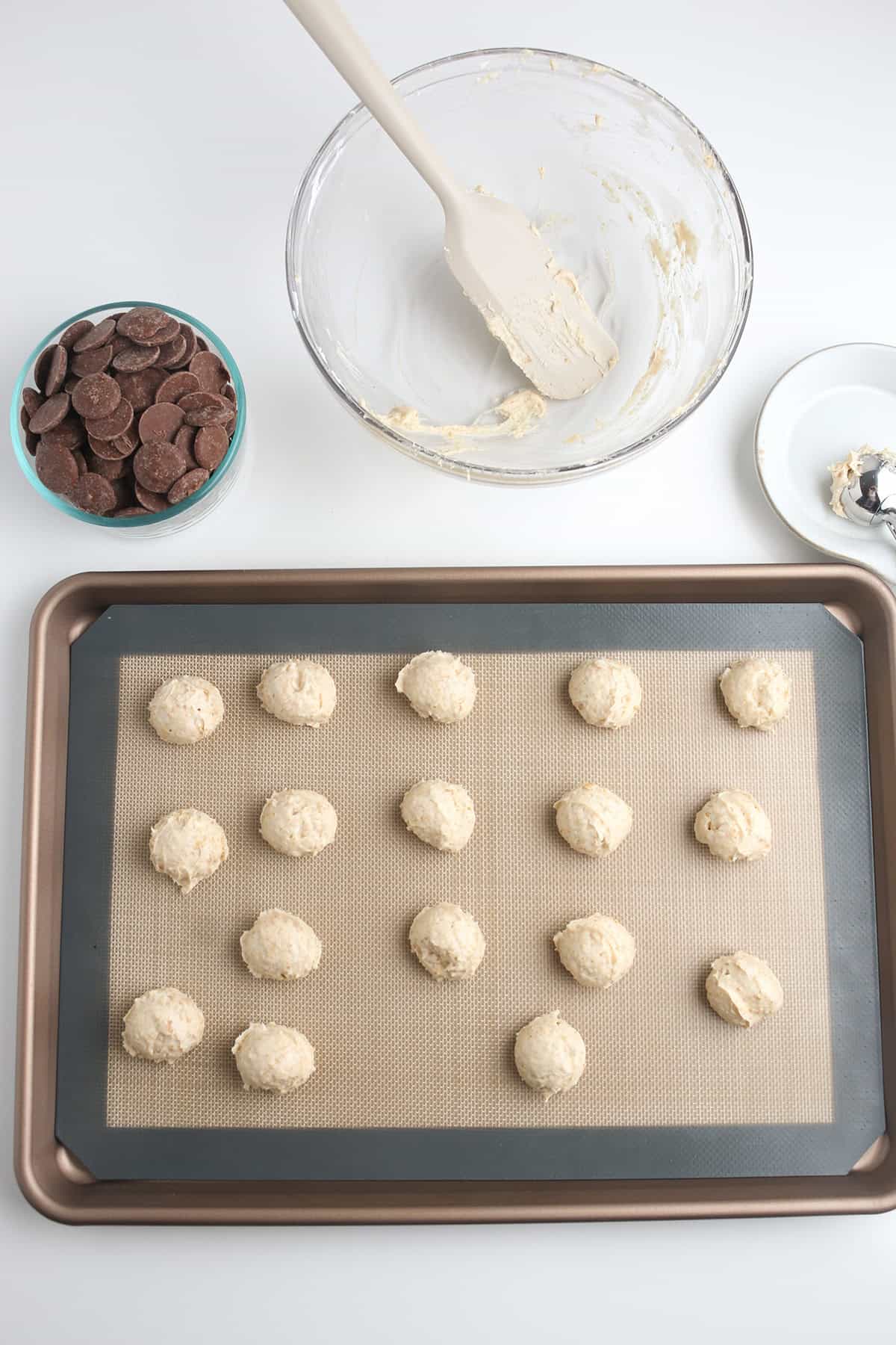 A baking tray with 16 cheesecake balls on a silicone mat, a bowl of chocolate pieces, and a mixing bowl with a spatula are arranged on a white surface.
