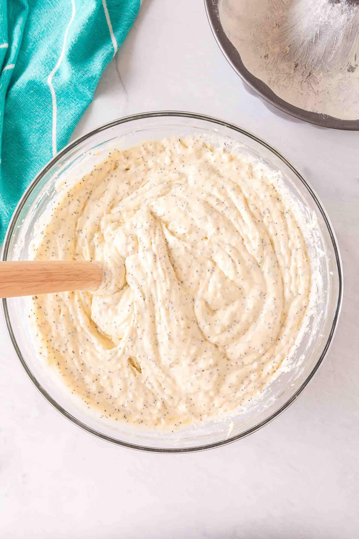 A glass bowl filled with Lemon Poppy Seed Bundt Cake batter being mixed with a wooden spatula, next to a green towel and a pie pan.