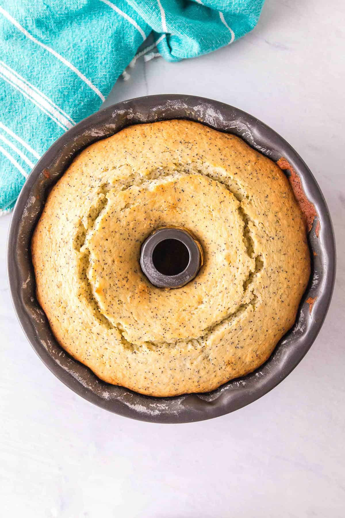 A baked Lemon Poppy Seed Bundt Cake sits in a round bundt pan on a white surface, accompanied by a teal striped kitchen towel.