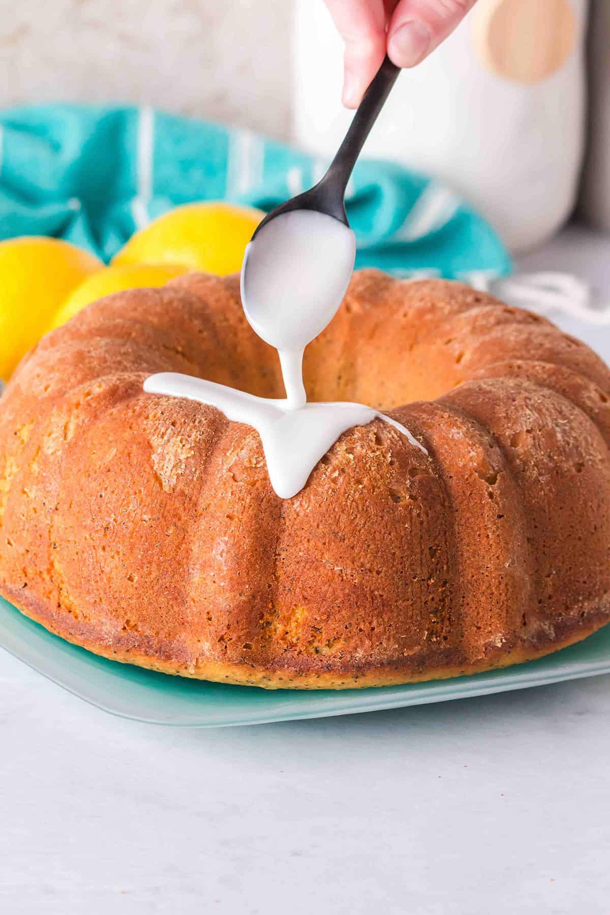 A hand pours white icing from a spoon onto a Lemon Poppy Seed Bundt Cake on a green plate, with lemons and a blue towel in the background.
