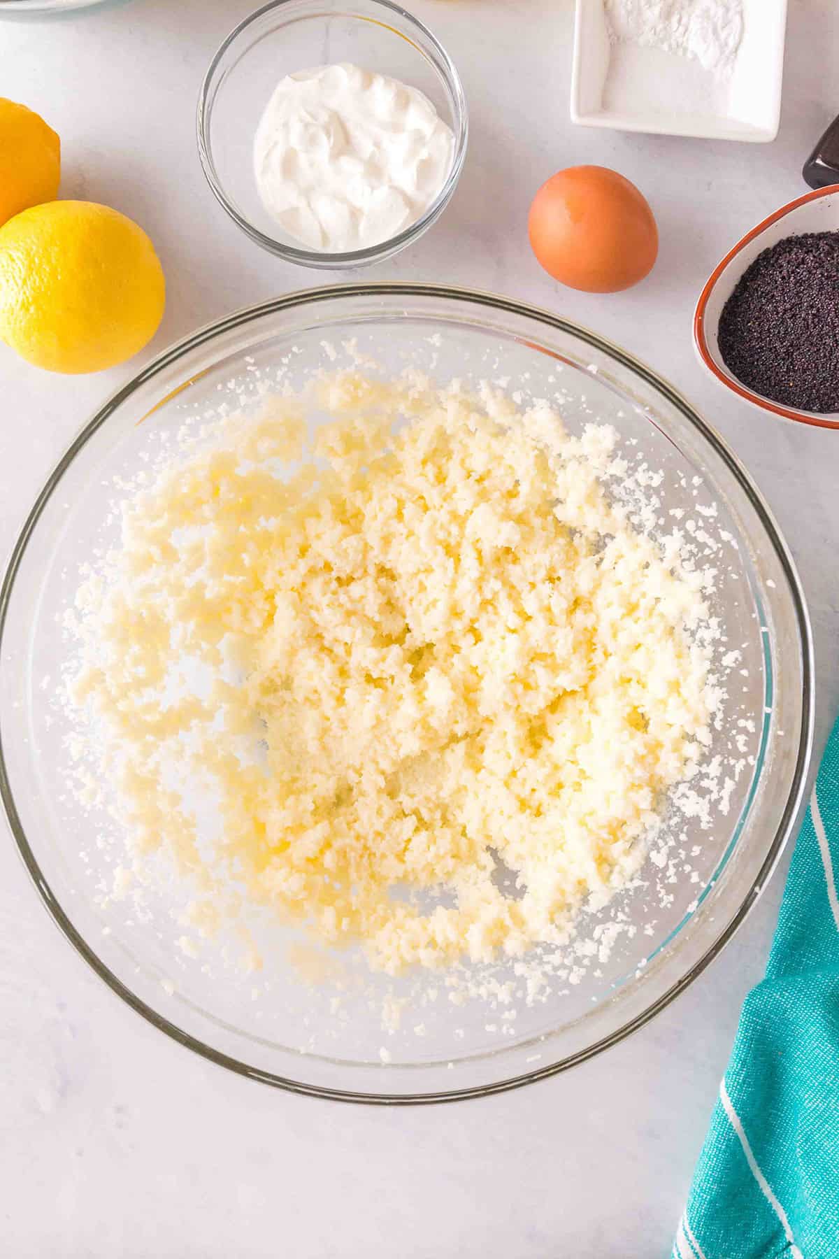 A glass bowl with creamed butter and sugar sits on a counter, surrounded by ingredients for Lemon Poppy Seed Bundt Cake: a lemon, an egg, a bowl of yogurt, poppy seeds, and flour.