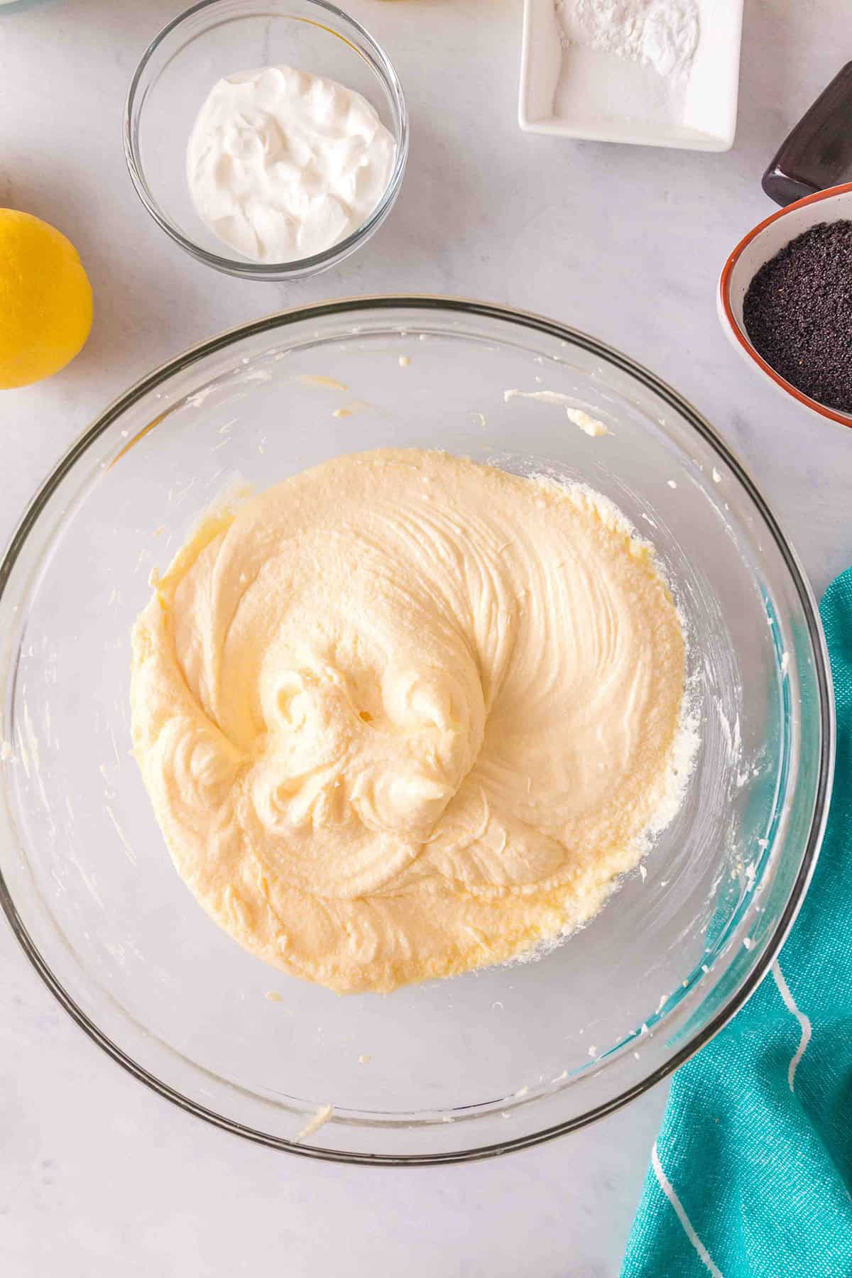 A glass bowl with creamed batter sits on a countertop, surrounded by small bowls of sour cream, poppy seeds, flour, and a lemon&mdash;perfect ingredients for a Lemon Poppy Seed Bundt Cake.