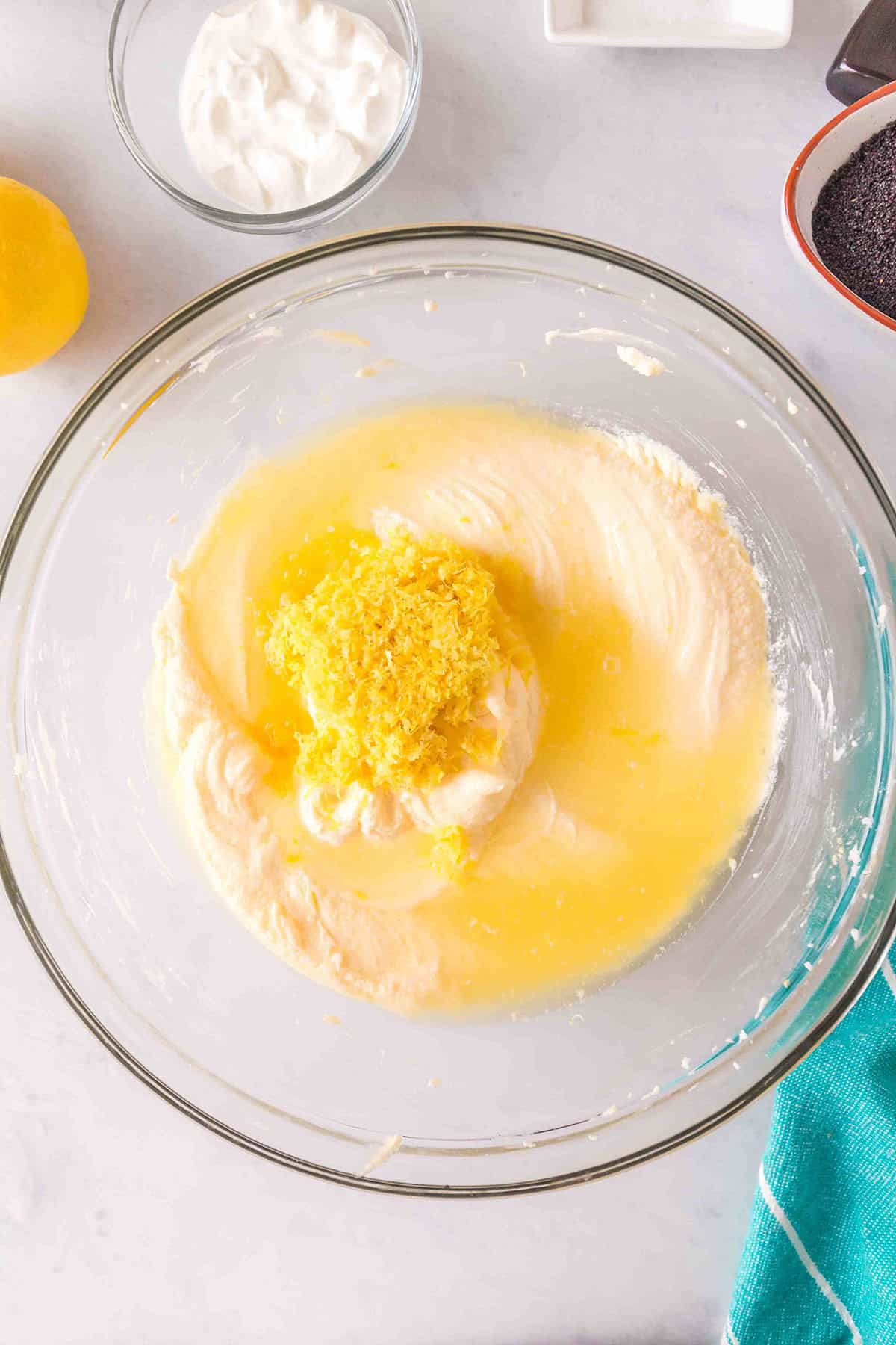 A glass bowl containing batter with lemon zest and juice on top, surrounded by a lemon, sour cream, poppy seeds, and a teal towel&mdash;perfect ingredients for a delicious Lemon Poppy Seed Bundt Cake.