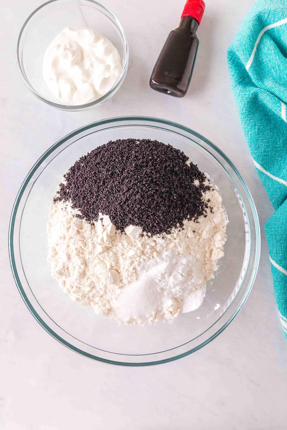 A glass bowl with flour, sugar, baking powder, and poppy seeds&mdash;perfect for making Lemon Poppy Seed Bundt Cake&mdash;plus a small bowl of sour cream, vanilla extract, and a teal towel on a white surface.