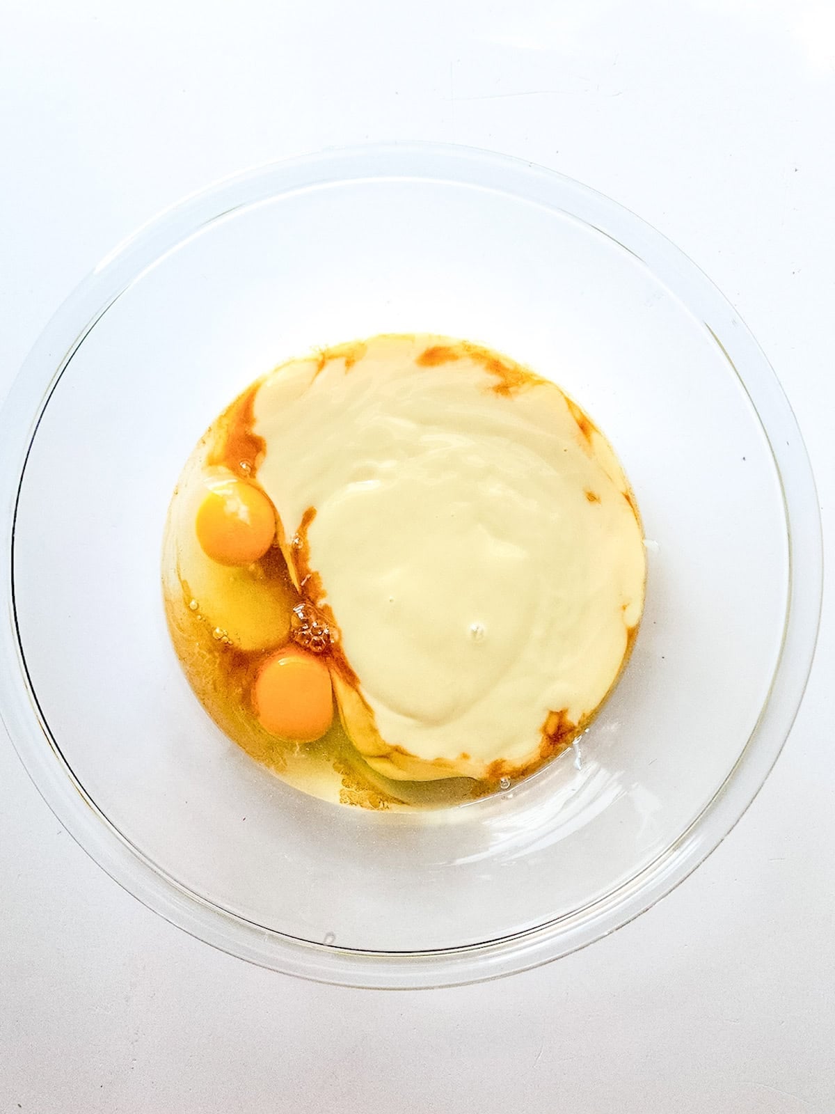 A clear glass bowl containing two raw eggs, condensed milk, and vanilla extract&mdash;ingredients ready to be mixed for delicious condensed milk brownies&mdash;viewed from above on a white background.