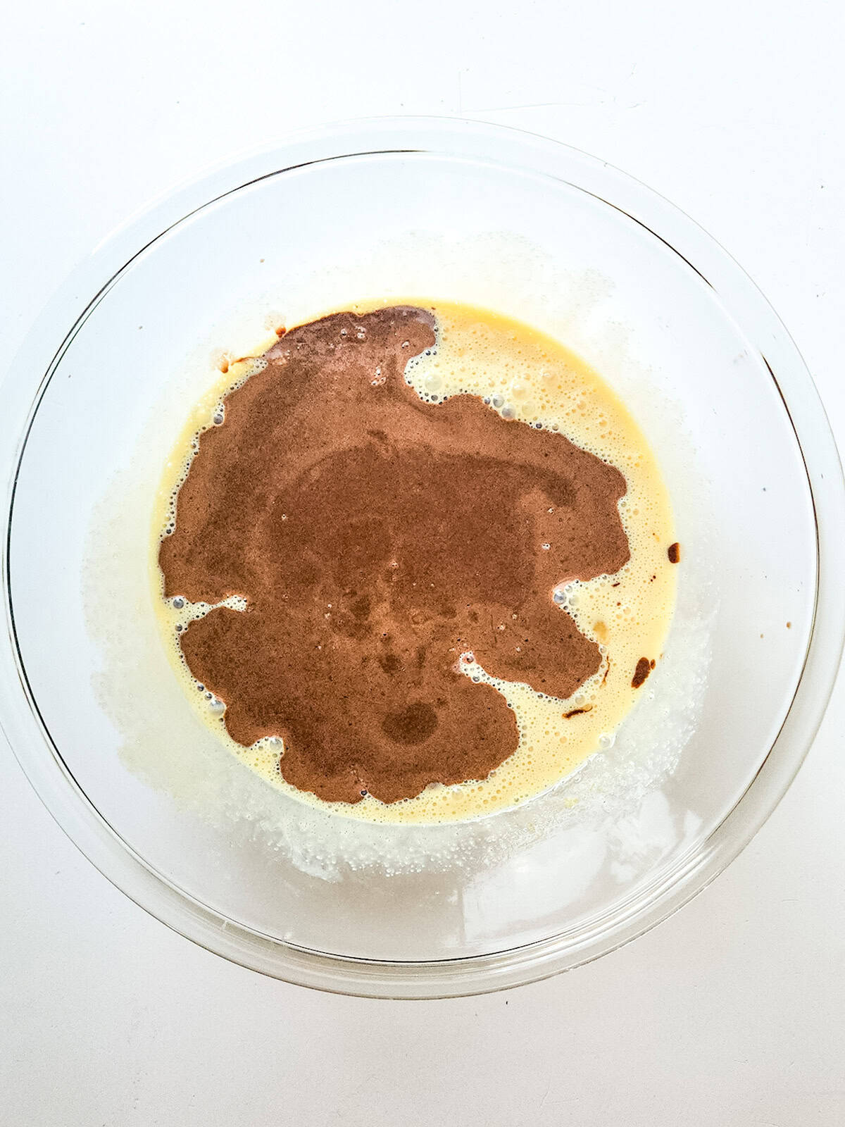 A glass bowl containing a mixture of light yellow batter with cocoa or chocolate swirled on top, viewed from above&mdash;perfect for preparing rich condensed milk brownies.