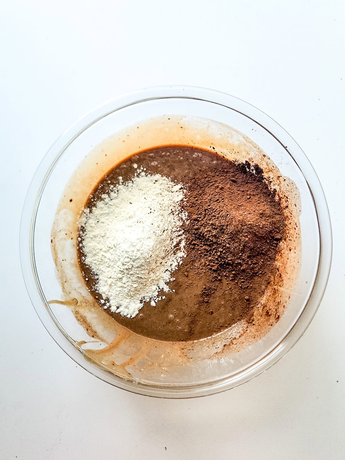A glass bowl with chocolate batter, ready for condensed milk brownies, topped with a mound of flour on one side and cocoa powder on the other, viewed from above.