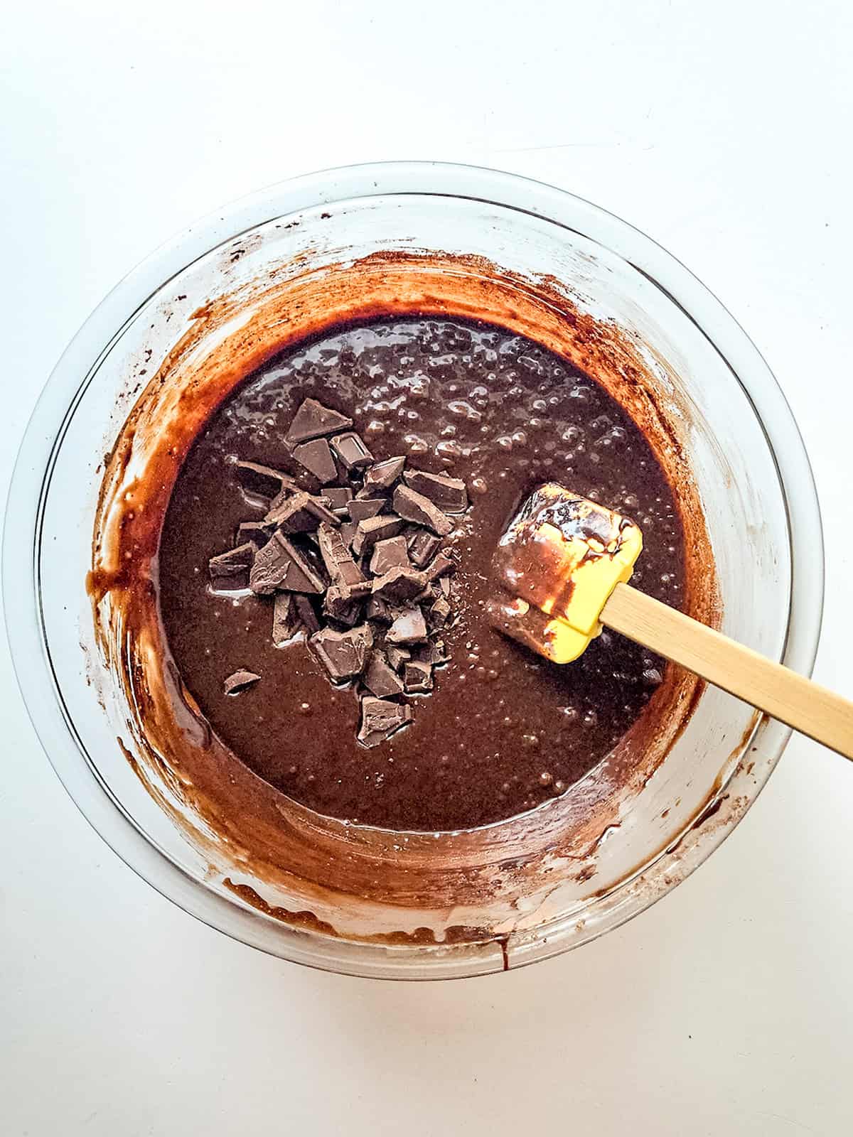 A glass bowl filled with sweetened condensed milk brownies batter, topped with chopped chocolate pieces, and a spatula with batter on it resting inside the bowl.