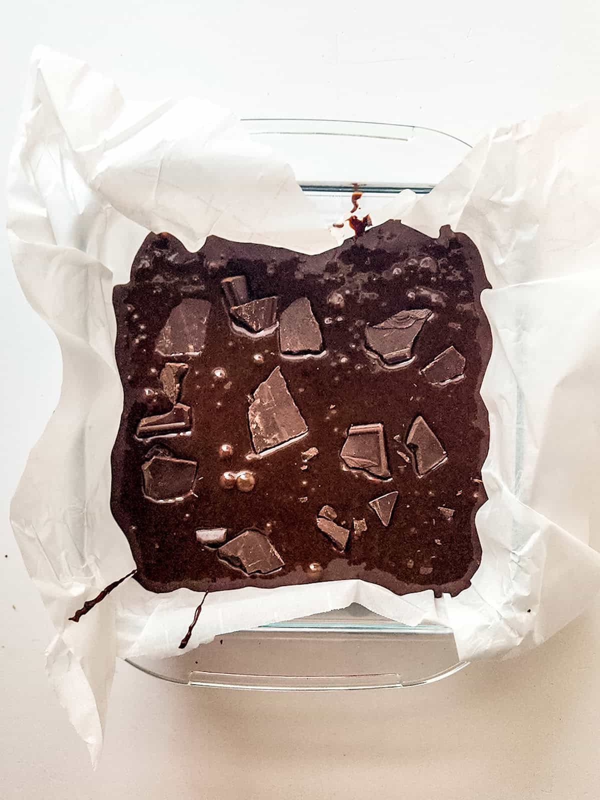 A square glass baking dish lined with parchment paper holds unbaked sweetened condensed milk brownies batter topped with chunks of chocolate.