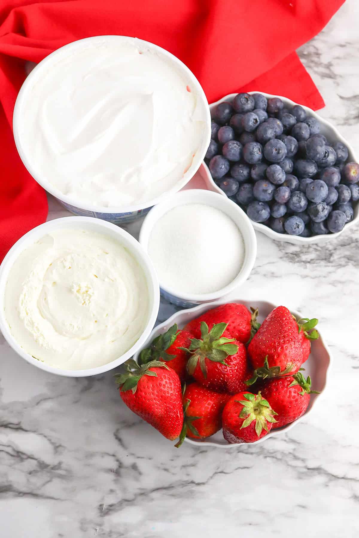 Bowls of Berry Cheesecake Salad with whipped cream, blueberries, sugar, cream cheese, and strawberries are arranged on a marble surface with a red cloth nearby.