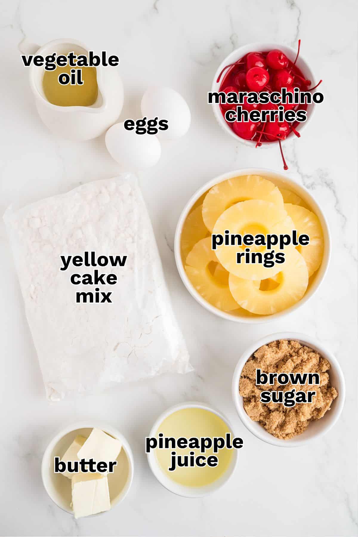 Baking ingredients for pineapple upside down cake on a white surface: yellow cake mix, eggs, vegetable oil, butter, pineapple juice, pineapple rings, brown sugar, and maraschino cherries.