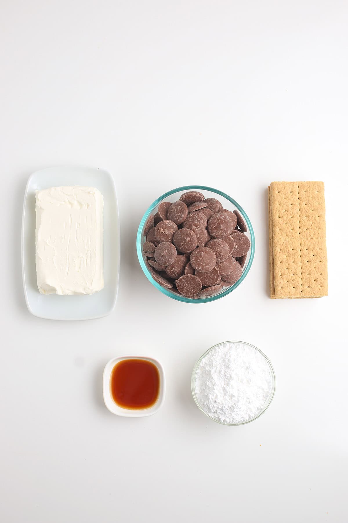 Five ingredients arranged on a white surface: cream cheese, chocolate wafers, graham crackers, vanilla extract, and powdered sugar&mdash;perfect for making delectable cheesecake truffles.