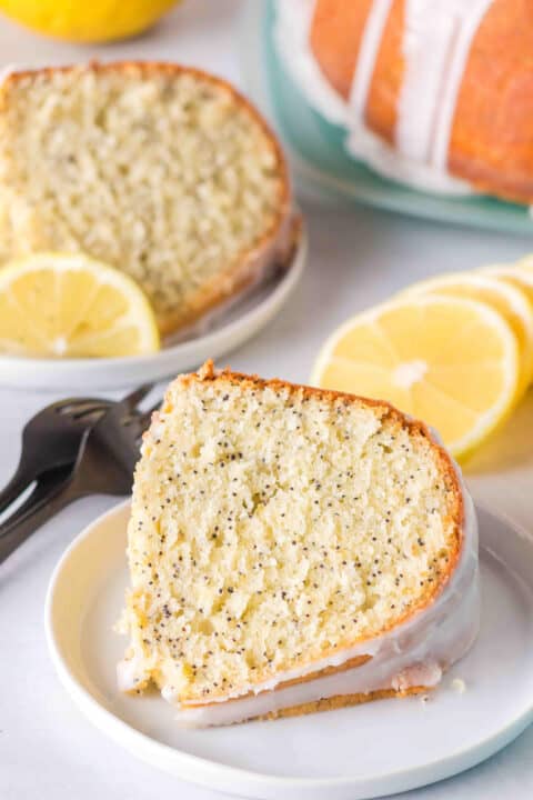 A slice of Lemon Poppy Seed Bundt Cake with glaze sits on a white plate, garnished with a lemon slice; the whole cake and extra lemon slices are displayed in the background.