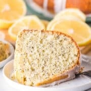 A slice of Lemon Poppy Seed Bundt Cake with icing rests on a white plate, garnished with fresh lemon slices in the background.