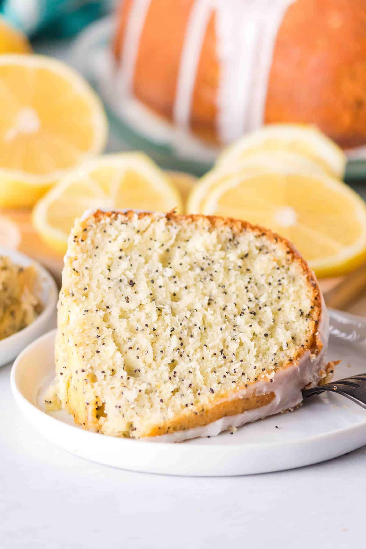 A slice of Lemon Poppy Seed Bundt Cake with icing rests on a white plate, garnished with fresh lemon slices in the background.